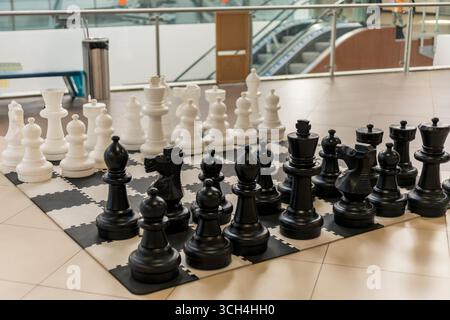 A large chess set features black and white pieces on a black and white checkered board. Players of all ages gather around, enjoying the outdoor Stock Photo
