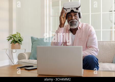 Senior man using VR headset and laptop at home, exploring virtual reality Stock Photo