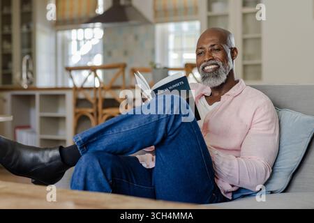 Smiling senior man reading book on sofa at home, enjoying leisure time Stock Photo