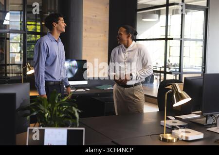 Two businessmen in modern office discussing work, smiling and standing near desks Stock Photo