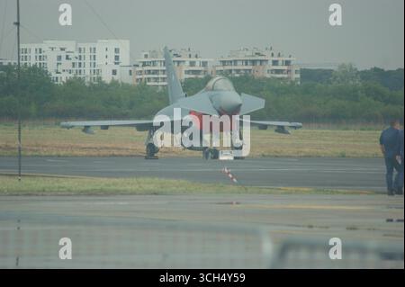 A Eurofighter Typhoon jet is parked on a tarmac, set against a backdrop of modern urban buildings, representing military technology and urban life. Stock Photo