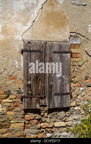 Weathered wooden shutters, reinforced with rusty metal, cover a window in a crumbling brick and plaster wall. The cracked texture hints at neglect and Stock Photo