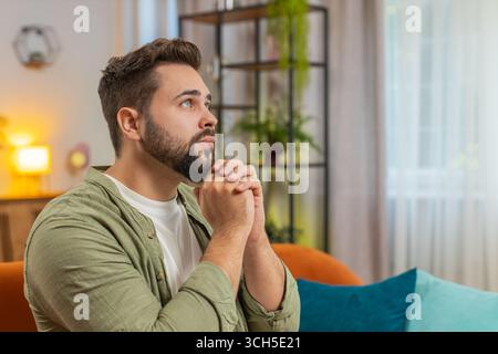 Tensed young man sitting on sofa at home folding palms together pray ask for help, thinks feel stressed looks desperate by life problems. Caucasian guy praying asking for help sitting on sofa at home. Stock Photo