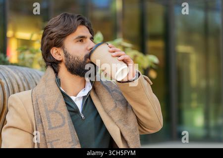 Happy Indian young man enjoying morning coffee hot drink and smiling outdoors. Relaxing, taking a break. Lebanese guy sitting on urban downtown city street drinking coffee to go. Town lifestyles Stock Photo