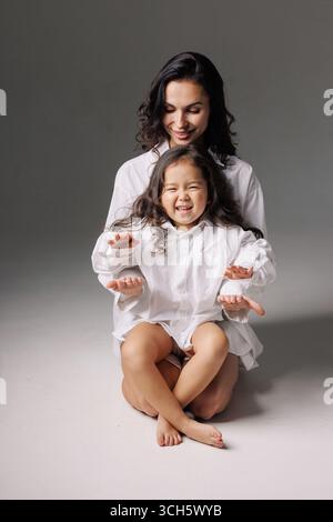 Mother and daughter smiling while sitting together in matching outfits playing hands in minimal studio setting Stock Photo