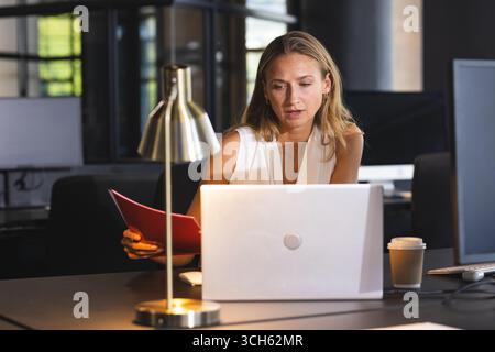Businesswoman reviewing documents and working on laptop in modern office at night Stock Photo