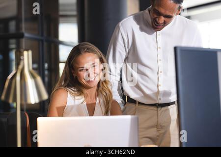 Colleagues collaborating on laptop in modern office, smiling and discussing project Stock Photo