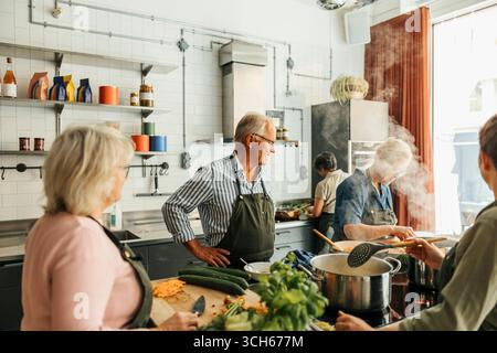 Elderly man looking at female friend preparing food while standing with hand on hip during cooking class in commercial k Stock Photo