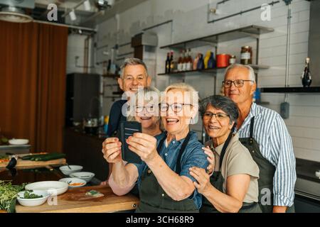 Happy elderly woman taking selfie with friends in commercial kitchen Stock Photo