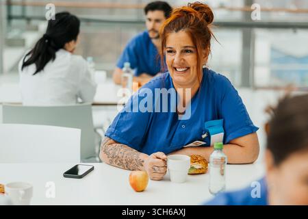 Smiling mature female nurse having breakfast while sitting at table in hospital cafeteria Stock Photo