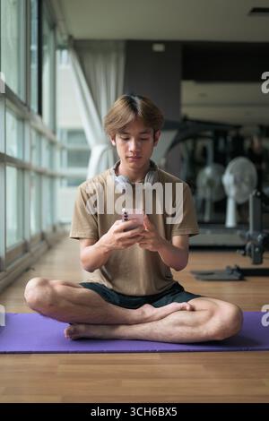 Young Thai Asian man with headphones sits cross-legged on a yoga mat near a large window in a fitness studio, focused on his smartphone with wellness, Stock Photo
