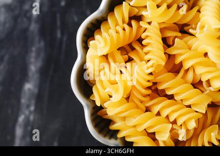 Spiral fusilli pasta in ceramic bowl. Closeup of raw noodles on marble table. Italian food ingredient for cooking. Yellow macaroni texture. Stock Photo