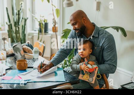 Businessman talking on smart phone and taking notes while sitting with son at home Stock Photo