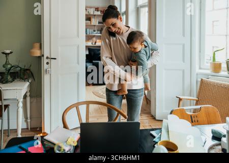 Smiling mother carrying son while talking on smart phone at home Stock Photo