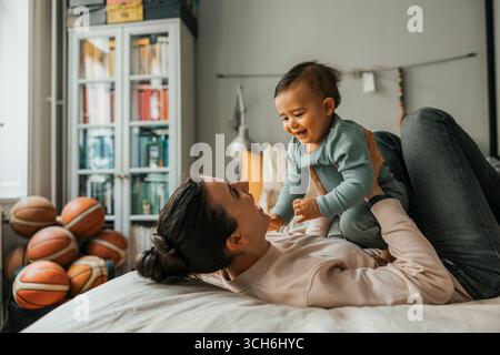 Happy woman playing with baby boy while lying on bed at home Stock Photo