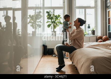 Mother playing with baby boy while sitting on bed at home Stock Photo