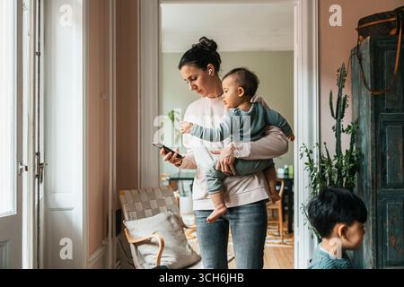 Woman wearing wireless in-ear headphones standing with baby boy while using smart phone at home Stock Photo