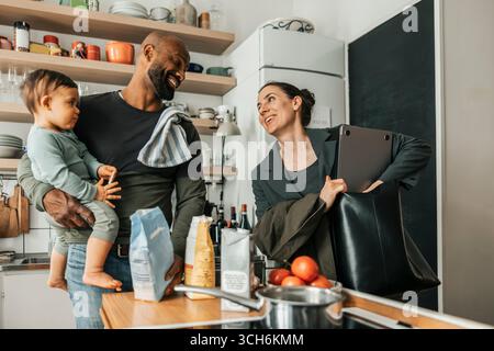 Smiling woman keeping laptop in bag while talking with man carrying son at home Stock Photo