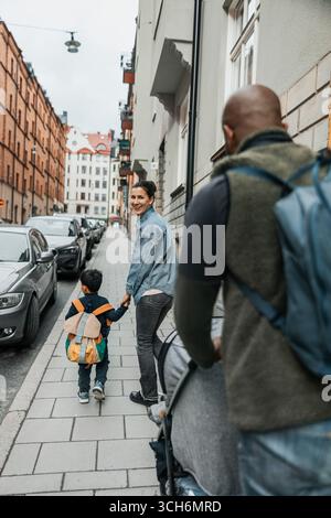 Happy woman holding hands of son looking over shoulder while walking on sidewalk Stock Photo
