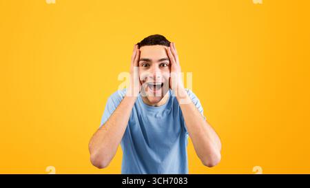Unbelievable promo. Handsome young man holding his head in shock on orange studio background Stock Photo