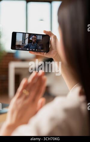 Young businesswoman doing hello hand gesture at black male colleague during online meeting on smartphone. Remote professionals collaborate on strategy during virtual call in home office. Stock Photo