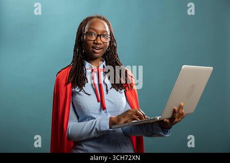 African american woman wearing a red cape and holding laptop with wide eyes, showing an expressive reaction of surprise. Black female model looking excited, as if receiving unexpected positive news. Stock Photo