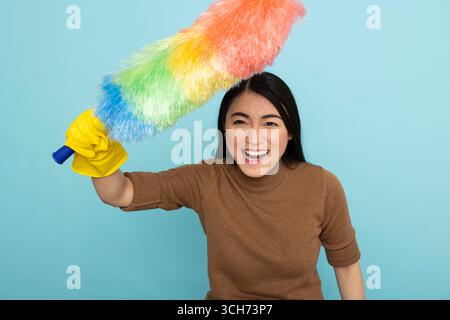 Young asian housekeeper with smile on face, excitedly holding feather duster, ready to tackle chores. Enthusiastic female cleaner prepared for professional household cleaning tasks. Stock Photo