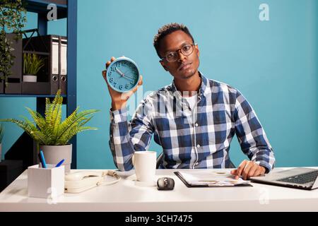 Portrait of african american freelancer sitting with a clipboard at desk and holding an alarm clock. Young business analyst handling paperwork and presenting the time with an analogue clock in hand. Stock Photo