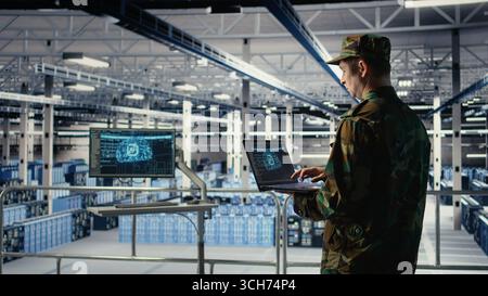 Soldier in engineering role uses AI application on laptop to manage server hub systems. Army commander reviews secret intelligence information using artificial intelligence in data center, camera A Stock Photo