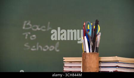 A stack of books with a pencil holder filled with school supplies in front of a chalkboard with "Back to School" written on it. Stock Photo