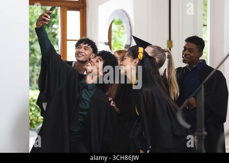 Graduating diverse students taking selfie in caps and gowns, celebrating together indoors, at home Stock Photo