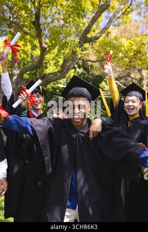 Celebrating graduation in garden, diverse students holding diplomas and wearing caps and gowns Stock Photo
