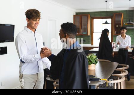 Teenage diverse graduates celebrating at home, shaking hands and smiling with diverse friends Stock Photo