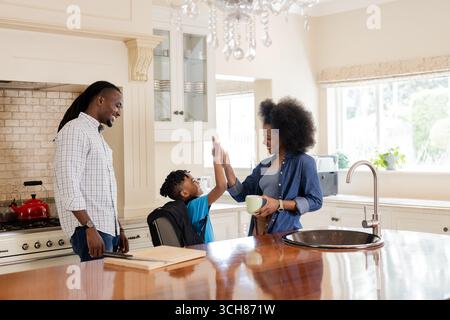 Family enjoying morning routine in kitchen, mother and son sharing high five Stock Photo