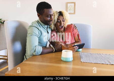 Sitting diverse couple using smartphone at wooden dining table at home, with ceramic mug Stock Photo