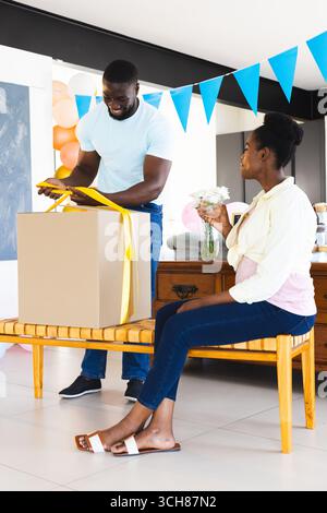 Man opening gift box while woman holding flowers, celebrating at home Stock Photo