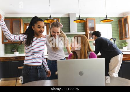 Teenagers celebrating success while using laptop in modern kitchen together Stock Photo