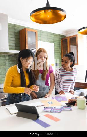 In kitchen, teen girls using tablet and colorful papers for creative project Stock Photo