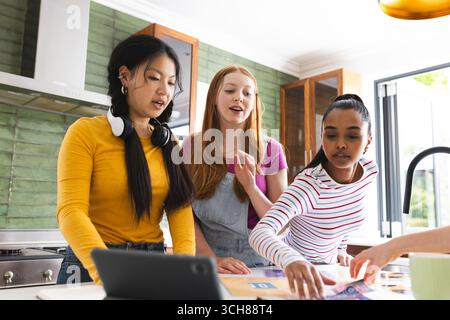 Teen girls collaborating on school project in kitchen, using tablet and papers Stock Photo