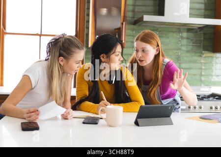 Teen girls collaborating on school project in kitchen, using tablet and notes Stock Photo