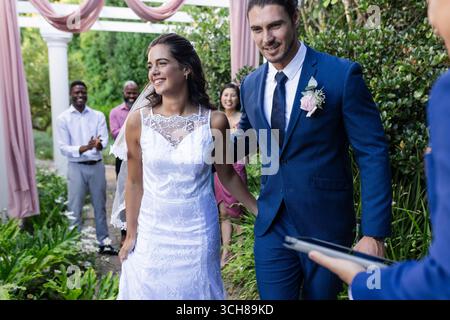 Bride and groom walking joyfully down garden aisle, guests applauding happily Stock Photo