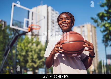 Young African American woman smiling while holding basketball on outdoor urban court. Stock Photo
