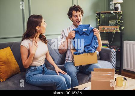A young caucasian couple joyfully unpacks a cardboard box at home, capturing the excitement of receiving a new shirt and enjoying domestic life togeth Stock Photo