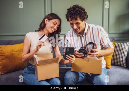 A happy young couple opens a cardboard package at home, smiling as they discover their purchase. She holds a credit card while he lifts the box, showc Stock Photo