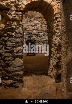Stone archway leading into medieval torture chamber with wooden cross and stool, creating eerie atmosphere. Stock Photo