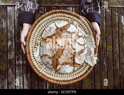In a wicker basket lined with cloth rests a rustic artisan loaf, its floury crust beautifully cracked. Hands hold it above a weathered wooden table Stock Photo