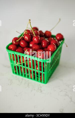Green basket of shiny red cherries on white marble countertop Stock Photo