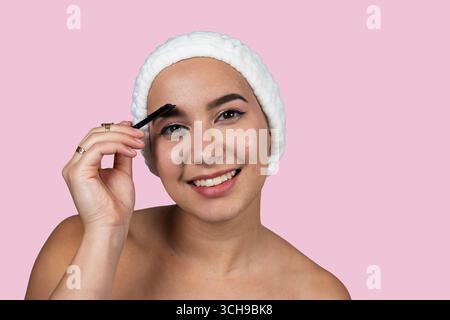 Beauty routine of a young woman combing her eyebrows with a brush after a shower, on a pink background Stock Photo