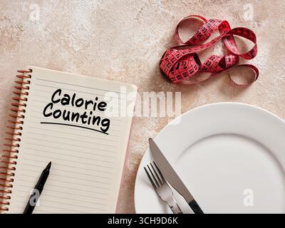 Notebook with calorie counting message, empty plate, cutlery and measuring tape suggesting a weight loss diet Stock Photo