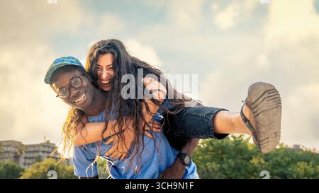 Happy multicultural couple having fun outdoors on a sunny summer day. Smiling young woman hugs man from behind, concept of diversity, friendship, love Stock Photo
